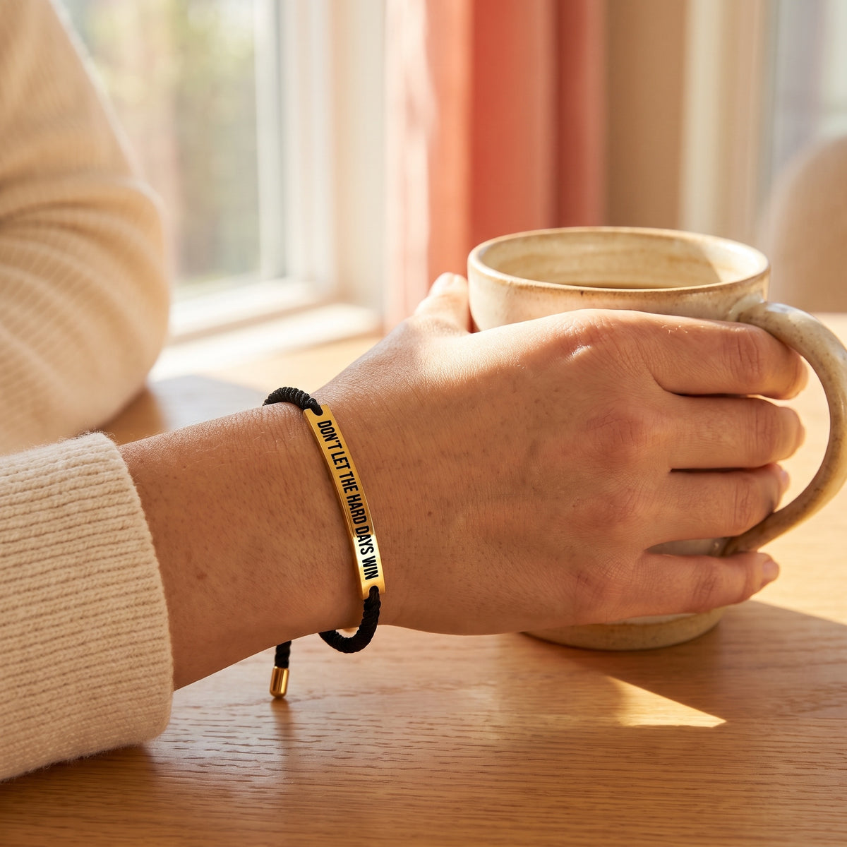 Woman holding a coffee mug wearing the black and gold Don't Let The Hard Days Win bracelet