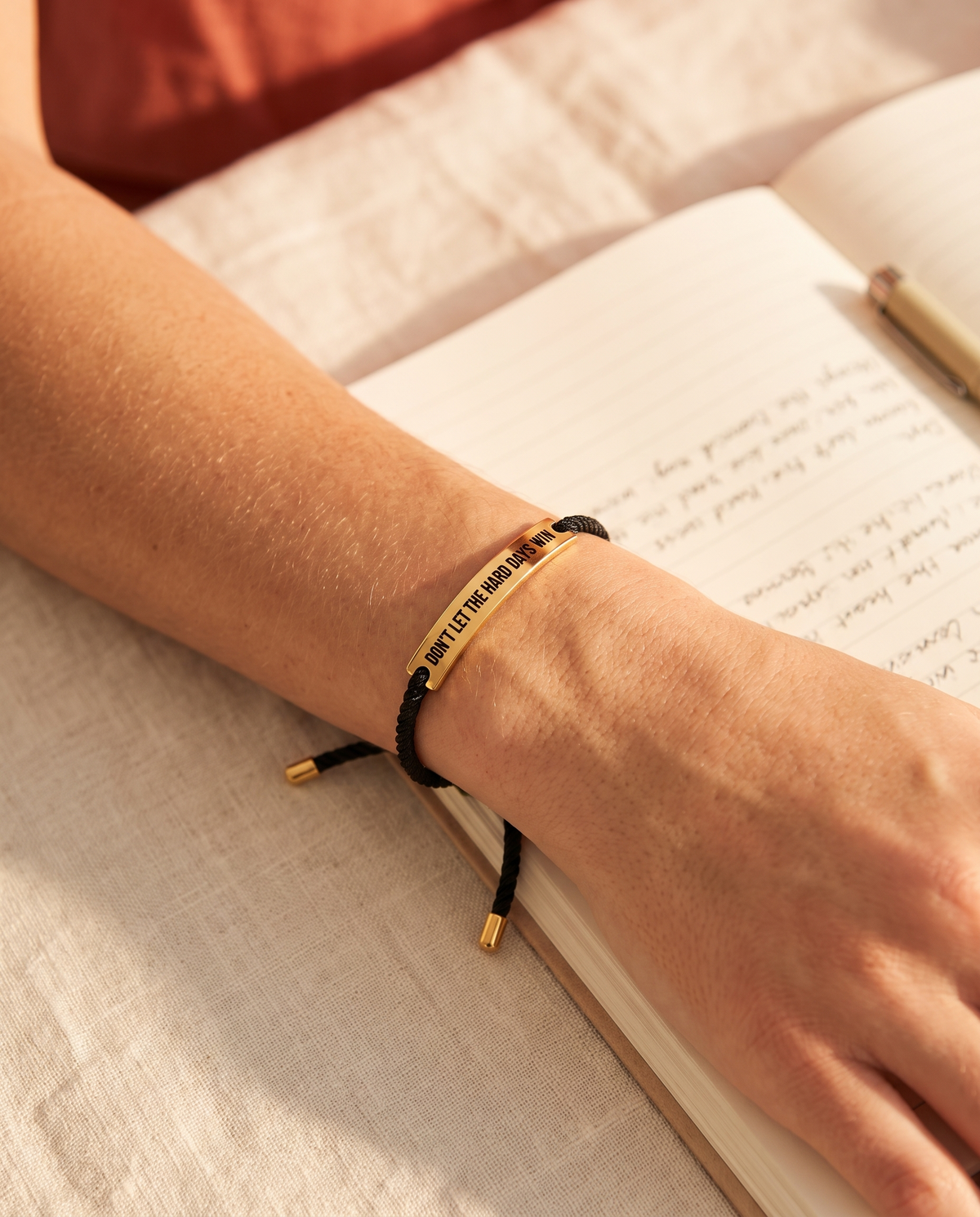 Wrist with the black and gold Don't Let The Hard Days Win bracelet resting on an open notebook